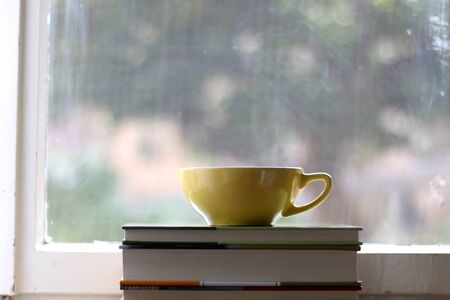 Tea in a yellow cup and stack of books on a rustic window sill. Selective focus, view of garden in the background.の写真素材