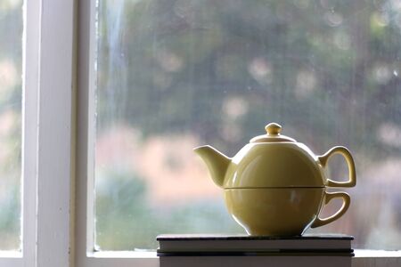 Yellow teapot and stack of books on a rustic window. Selective focus, view of garden in the background.の写真素材