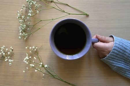 Unrecognizable person in a cozy sweater holding a cup of tea, with gypsophila flowers on wooden...