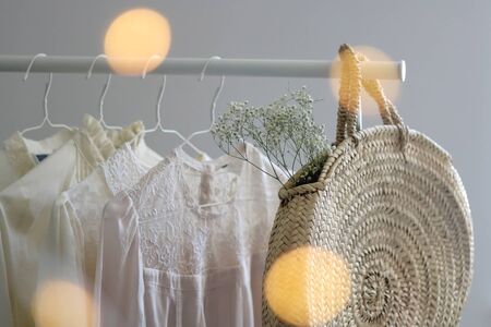Fashionable wicker bag and white and neutral blouses on a clothing rack. Selective focus, yellow bokeh lights in the foreground.の写真素材