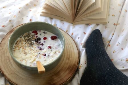 Unrecognizable person in fuzzy socks eating breakfast bowl in bed, with open book. Selective focus.の写真素材