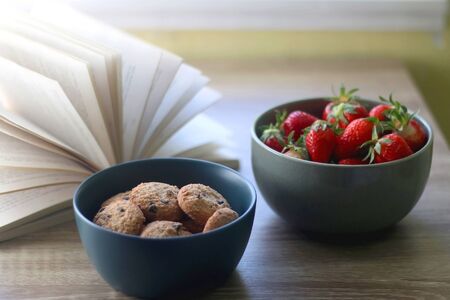 Bowl of chocolate chip cookies, bowl of strawberries and an open book on a table. Selective focus.の写真素材