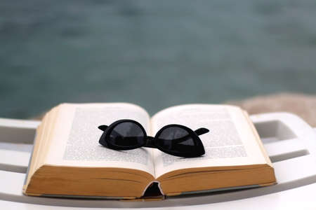 Open book and fashionable sunglasses on a deck chair by the water. Selective focus.の写真素材