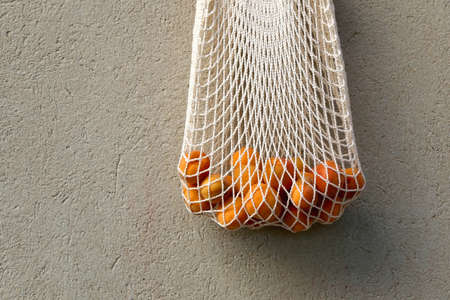 Reusable mesh bag filled with fresh apricots, hanging on a concrete wall.