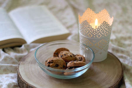 Wooden tray with bowl of chocolate chip cookies and lit candle on a bed. Open book in the background. Selective focus.の写真素材