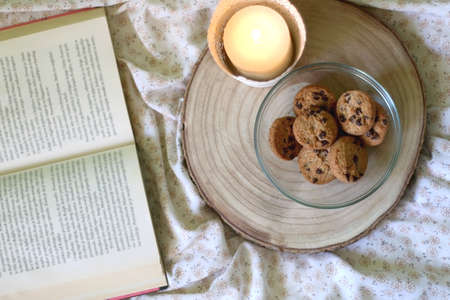 Wooden tray with bowl of chocolate chip cookies and lit candle on a bed. Open book in the background. Top view.の写真素材