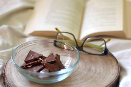 Bowl of chocolate, reading glasses and open book on a bed. Selective focus.の写真素材
