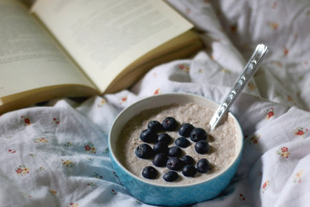 Bowl of porridge with cinnamon and blueberry topping, and open book on a bed. Selective focus.の写真素材