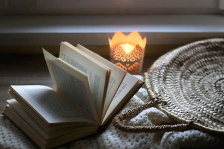 Open book, woven basket, crochet blanket and lit candle. Cozy details at home. Selective focus.の写真素材