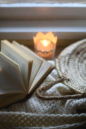 Open book, woven basket, crochet blanket and lit candle. Cozy details at home. Selective focus.の写真素材