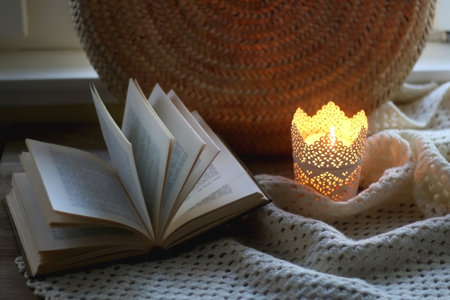 Open book, woven basket, crochet blanket and lit candle. Cozy details at home. Selective focus.の写真素材