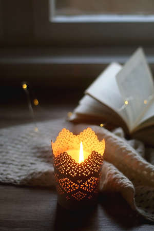 Lit candle, crochet blanket, open book and bokeh lights. Cozy home edtails. Selective focus.