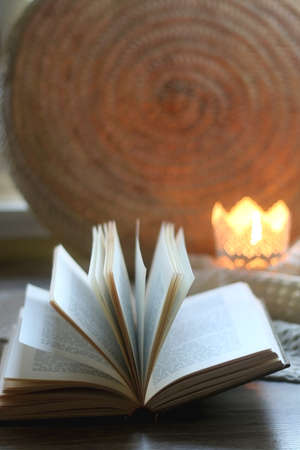 Open book, woven basket, crochet blanket and lit candle. Cozy details at home. Selective focus.