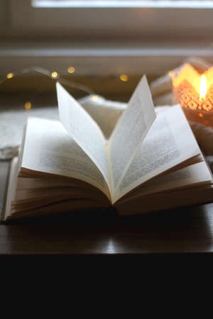 Open book, lit candle, crochet blanket and bokeh lights. Cozy home edtails. Selective focus.の写真素材