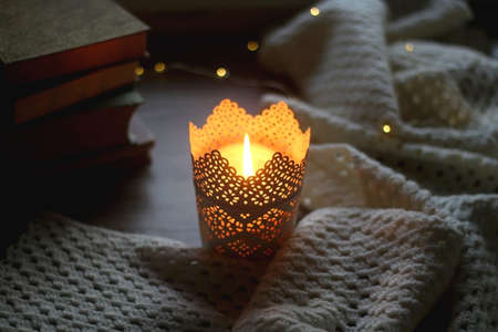 Candle holder with lit candle, soft knitted blanket, stack of books and bokeh lights at home....