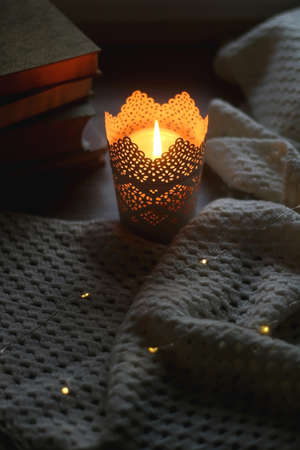 Candle holder with lit candle, soft knitted blanket, stack of books and bokeh lights at home. Selective focus.の写真素材