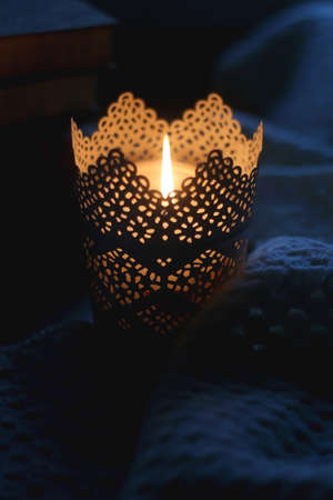 Candle holder with lit candle, soft knitted blanket and stack of books at home. Selective focus.