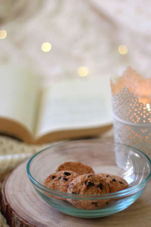 Bowl of chocolate chip cookies, lit candle and open book on a bed. Selective focus.