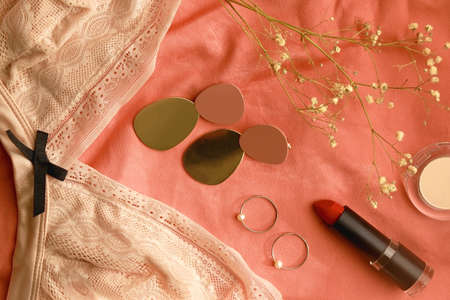 Lace bralette, make up products, earrings, rings and flowers on pink background. Top view.