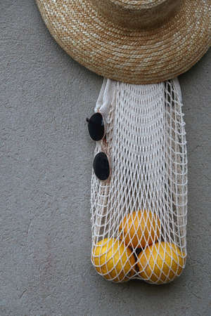 Reusable mesh bag filled with lemons, round sunglasses and straw hat, hanging on the concrete wall.の写真素材