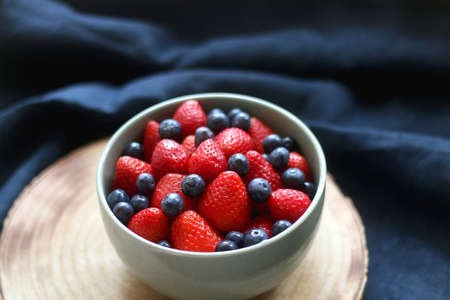 Bowl of blueberries and strawberries on wooden tray. Selective focus, dark textile background.の写真素材
