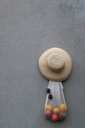 Reusable mesh bag filled with lemons, round sunglasses and straw hat, hanging on the concrete wall.
