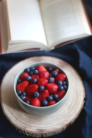 Bowl of blueberries and strawberries on wooden tray and open book. Selective focus, dark textile...