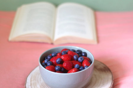 Bowl of blueberries and strawberries and open book on a bed. Selective focus.