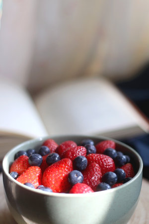 Bowl of blueberries and strawberries on wooden tray and open book. Selective focus, dark textile background.の写真素材