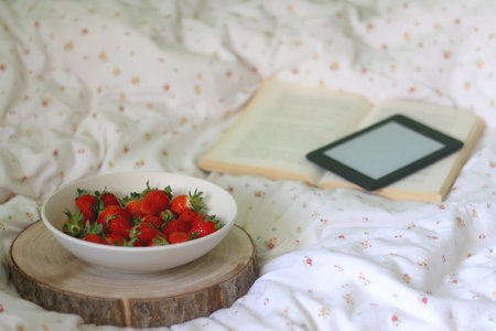 Bowl of strawberries, open book and e-reader on a bed. Selective focus.の写真素材