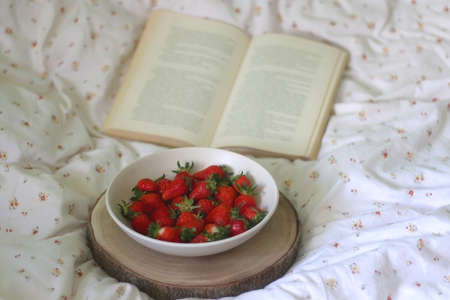 Bowl of strawberries and open book on a bed. Selective focus.の写真素材