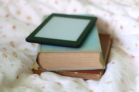 Stack of old hardcover books and digital e-reader on a bed. Selective focus.の写真素材