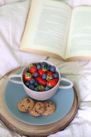Bowl of blueberries and strawberries, chocolate chip cookies and open book on a bed. Selective focus.の写真素材