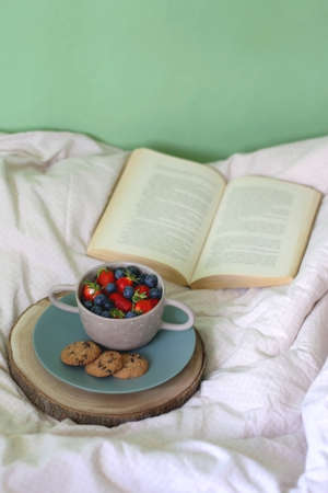 Bowl of blueberries and strawberries, chocolate chip cookies and open book on a bed. Selective...