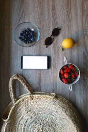 Straw bag, phone with white empty screen, sunglasses and various fruit on wooden background. Flat lay.の写真素材