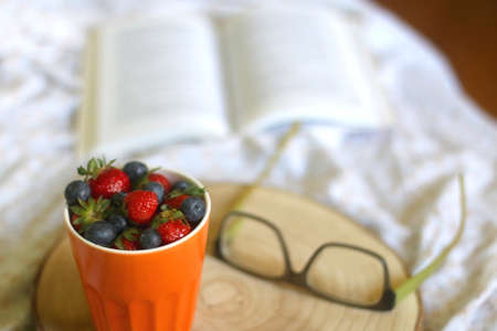 Cup of blueberries and strawberries, open book and reading glasses on a bed. Selective focus.の写真素材