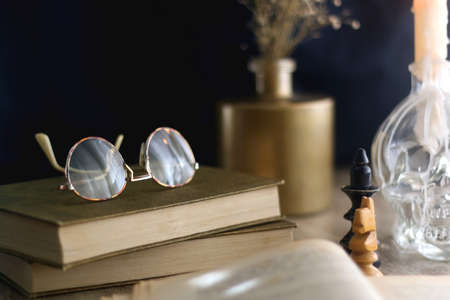 Old books, reading glasses, vintage chess pieces, lit candle and vase with gypsophila flowers. Dark academia concept. Selective focus.の写真素材