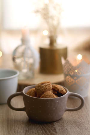 Bowl of cinnamon cookies, cup of warm drink, lit candles and vase with gypsophila flowers on the table. Selective focus, fairy lights in the background. Hygge at home.の写真素材