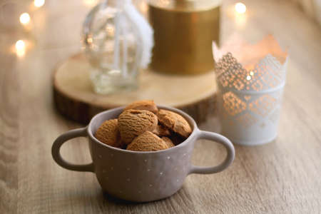 Bowl of cinnamon cookies, cup of warm drink, lit candles and vase with gypsophila flowers on the table. Selective focus, fairy lights in the background. Hygge at home.の写真素材