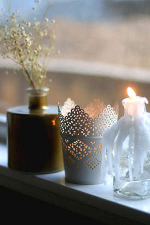 Candle holders with lit candles and vase with gypsophila flowers on a window sill. Hygge at home. Selective focus.の写真素材