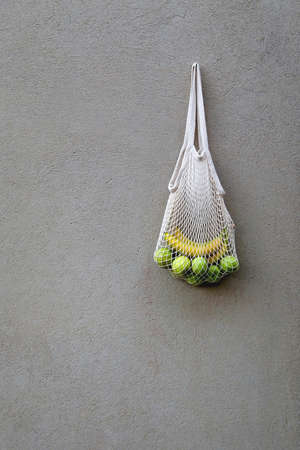 Crochet bag filled with green apples and bananas, hanging on a concrete wall.の写真素材