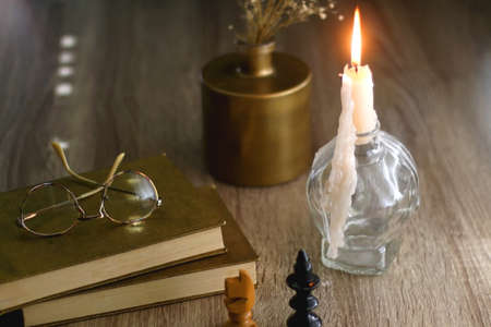 Old books, reading glasses, vintage chess pieces, lit candle and vase with gypsophila flowers. Dark academia concept. Selective focus.の写真素材
