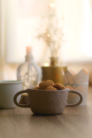 Bowl of cinnamon cookies, cup of warm drink, lit candles and vase with gypsophila flowers on the table. Selective focus, fairy lights in the background. Hygge at home.の写真素材