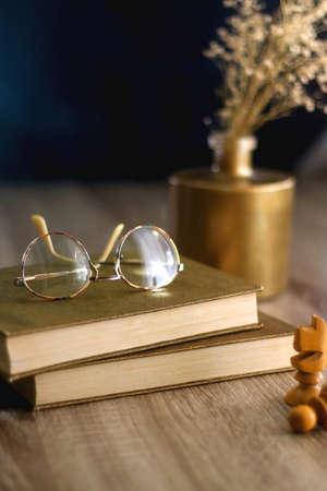 Old books, reading glasses, vintage chess pieces, lit candle and vase with gypsophila flowers. Dark...