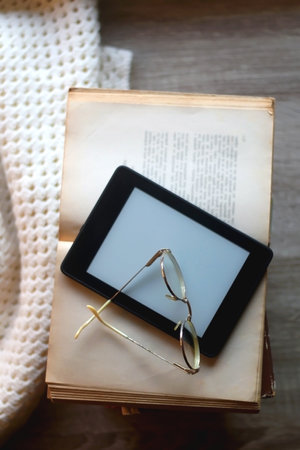 Stack of vintage books, e-reader, soft blanket and reading glasses. Hygge at home. Top view.