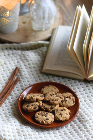 Plate of chocolate chip cookies, cinnamon sticks, soft blanket, open book and lit candles. Hygge at home. Selective focus.の写真素材