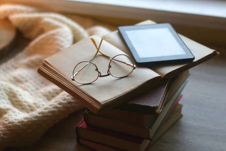 Stack of vintage books, e-reader, soft blanket and reading glasses. Hygge at home. Selective focus.の写真素材