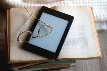Stack of vintage books, e-reader, soft blanket and reading glasses. Hygge at home. Selective focus.