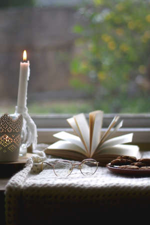 Plate of chocolate chip cookies, cinnamon sticks, soft blanket, open book and lit candles. Hygge at...