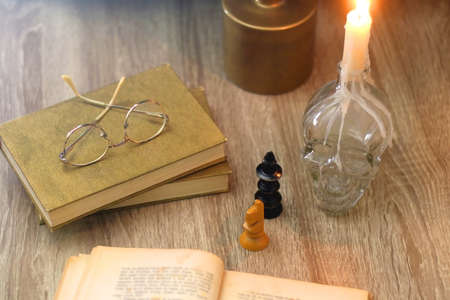 Old books, reading glasses, vintage chess pieces, lit candle and vase with gypsophila flowers. Dark...